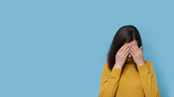 A frustrated female student in glasses and a sweater covering her eyes with her hands on a blue background, copy space. The girl is worried about her studies, love, relationships