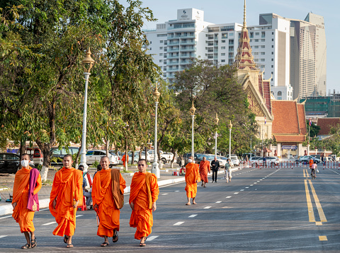 Row of Thai monks walking along people is   getting food donations  at  ceremony of Makha Bucha Day in Sangkhla Buri in northwest of Kanchanaburi province of Thailand in early morning close to sunset. People are wearing mostly traditional clothing and are standing in main street leading down to Mon Bridge and lake. People are waiting to give food donations to monks when they pass along street down to bridge.