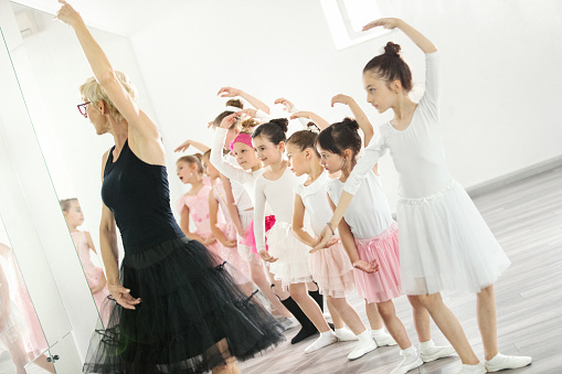 Closeup side view of elementary age girls practicing ballet moves with their teacher in front of a mirror at dance school.