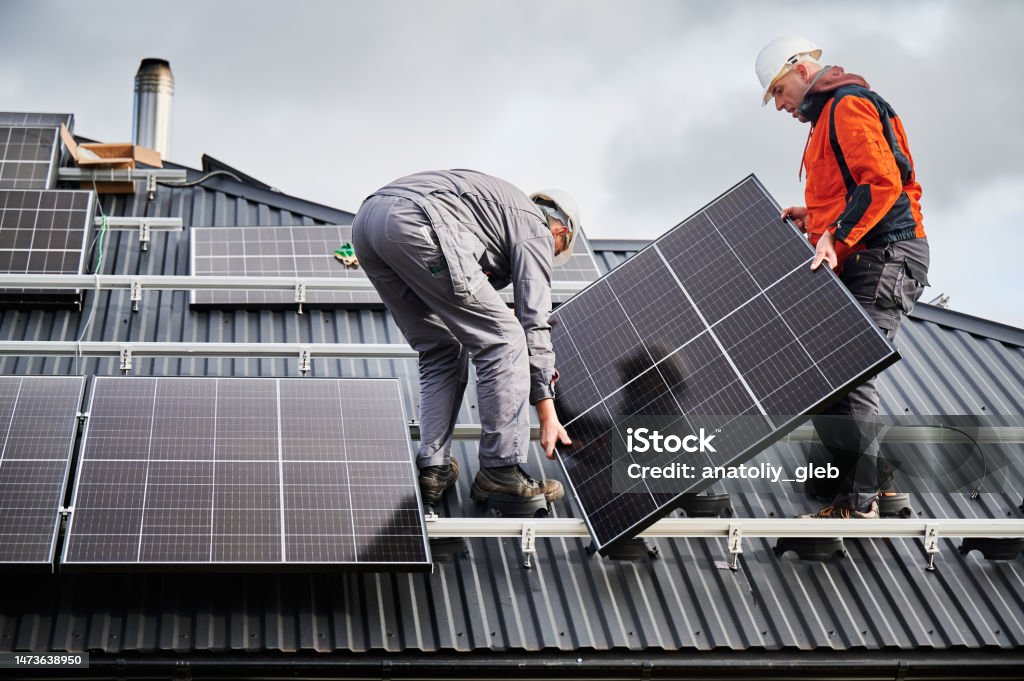 Technicians carrying photovoltaic solar module while installing solar panel system on roof of house Engineers building solar panel system on roof of house. Men workers in helmets carrying photovoltaic solar module outdoors. Concept of alternative and renewable energy. Solar Panel Stock Photo