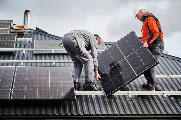 Engineers building solar panel system on roof of house. Men workers in helmets carrying photovoltaic solar module outdoors. Concept of alternative and renewable energy.