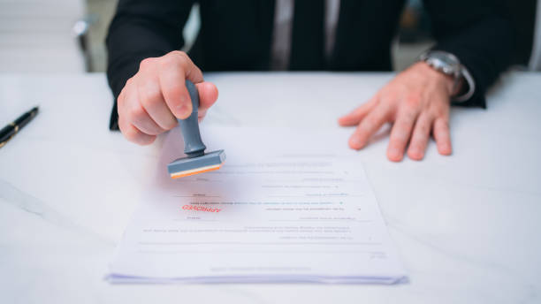 istock Businessman hand stamping an approved stamp on a text document at a table with a contract form paper in the background. 1473639108
