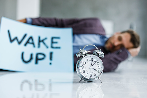 Close up of an alarm clock on the floor with a businessman in the background sleeping. Copy space.