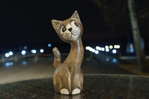 Handmade brown wooden cat figurine for living room home decoration. Lights of night city as background. Embankment of the sea. He looking at camera.