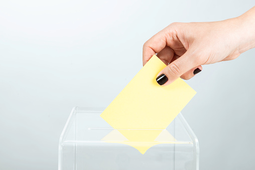 Human hand is inserting yellow envelope into ballot box.