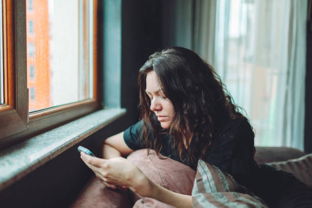 Mujer deprimida sentada en el sofá de su casa con el móvil - foto de stock