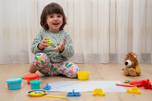 Female toddler holding a piece of yellow modelling clay while is looking at camera laughing.
