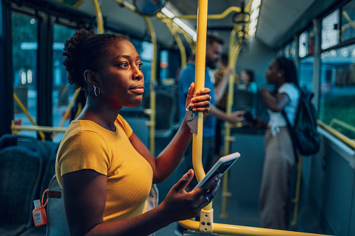 Young african american woman smiling while standing by herself on a bus and listening to music on a smartphone in the night. Female passenger using airpods and mobile phone in public transportation.
