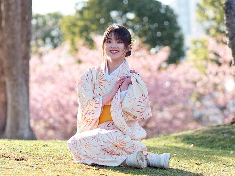 Portrait of beautiful young Chinese girl in Japanese kimono sitting on meadows with blossom cherry flowers background in spring garden, beauty, emotion, lifestyle, expression and people concept.