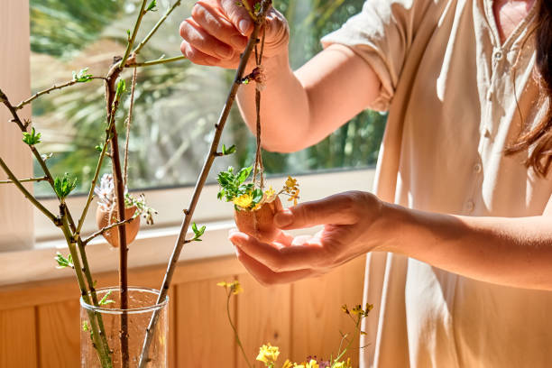 istock Easter home decoration. Woman's hands hanging egg shell with small wildflowers on spring twigs as easter decoration.Easter floral arrangement. 1473661942
