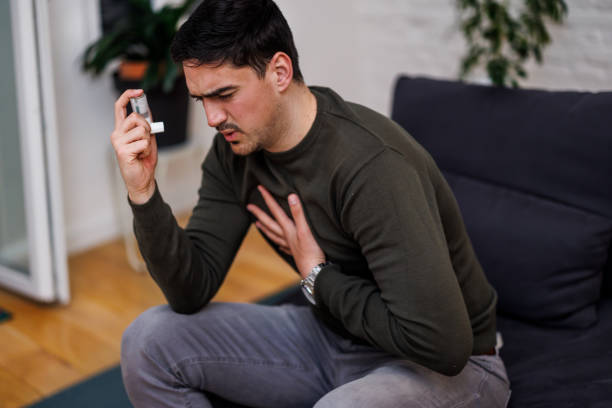 A young asthmatic man is suffering from chronic allergy and using an asthma inhaler. A young man is sitting on a sofa at home while suffering from a problem with breathing and using a smoke inhaler for asthma. asthma stock pictures, royalty-free photos & images