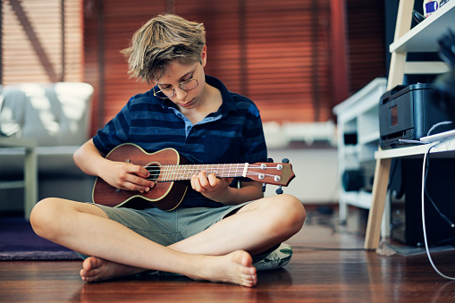 Cute little boy aged 11 playing ukulele at home. The boy is sitting on the floor and is very focused on playing.\nShot with Canon R5