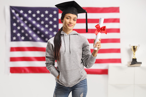 Female student with a graduation hat holding a diploma and posing in front of a USA flag