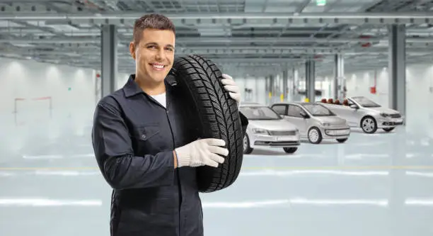 Auto mechanic worker carrying a tire on shoulder and smiling at camera inside a garage