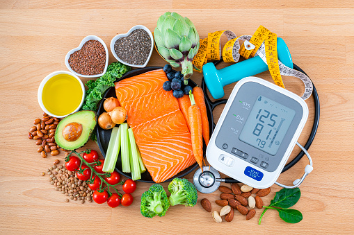 Heart care concept. Overhead view of a group of food that contains healthy nutrients for heart care, a blood pressure monitor, sthetoscope and dummbells shot on wooden table. The composition includes extra virgin olive oil, legumes, berries, nuts and seeds, salmon, broccoli, carrot, brown lentils, spinach, kale, artichoke and tomato among others.High resolution 42Mp studio digital capture taken with SONY A7rII and Zeiss Batis 40mm F2.0 CF lens