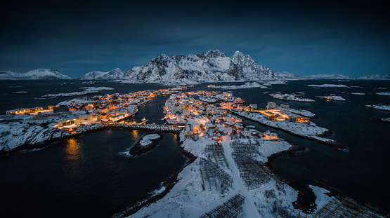 Aerial view of fishing village  Henningsvær in Nordland, Norway located on the Lofoten Islands archipelago. The village is illuminated at dusk and surrounded by cold water from the arctic ocean. Snowcapped mountains in the background. Nordland, Norway in Scandinavia , Europe.