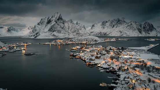 Aerial view of traditional Norwegian fishing Reine  at the Lofoten islands of Norway. The village is illuminated at dusk and surrounded by cold water from the arctic ocean. Snowcapped mountains in the background. Nordland, Norway in Scandinavia , Europe.