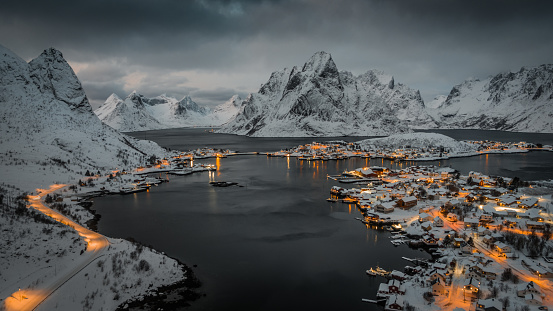 Aerial view of traditional Norwegian fishing Reine  at the Lofoten islands of Norway. The village is illuminated at dusk and surrounded by cold water from the arctic ocean. Snowcapped mountains in the background. Nordland, Norway in Scandinavia , Europe.