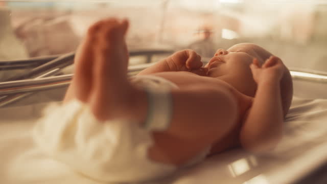 Cute Little Caucasian Newborn Baby Lying in Bassinet in a Maternity Hospital. Portrait of a Tiny Playful and Energetic Child with a Name ID Tag on the Leg. Warm Color Filter and Shading