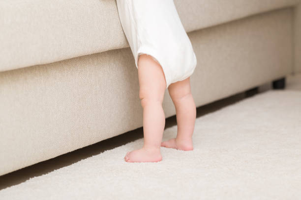 istock 8 months old baby in white bodysuit with barefoot standing on light soft carpet at beige sofa at living room. Ready to climb up on bed. Side back view. Closeup. 1473687730