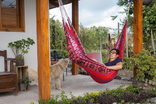 Latino man reclining in a red hammock working on his laptop with a dog accompanying him. High quality photo