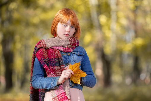 Sad pensive red-haired girl with a maple leaf in the autumn park.