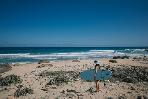 solo female Asian Chinese wave surfer standing on beach looking at sea carrying surfboard