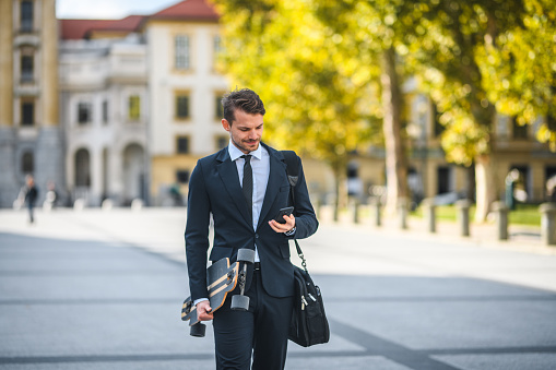 Handsome Caucasian male entrepreneur, carrying a longboard and a messenger bag, smart phone in hand, walking down the street of old town in Europe, city park in view. Dressed elegantly, early morning, slowly going to work. Focus on foreground. Looking away, medium full length image.