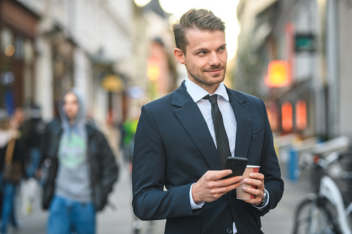 Waist up shot of handsome stock broker holding a coffee-to-go and a smart phone, on a busy city street. Looking away, blurred background.