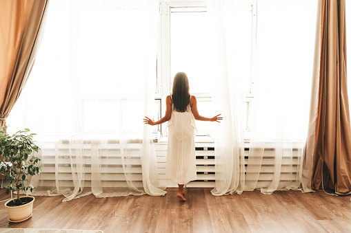 New day. Rear view of young brunette woman after waking up stands by large window in silk white gown and opening curtains with both hands, letting first morning rays of sun into bedroom at home