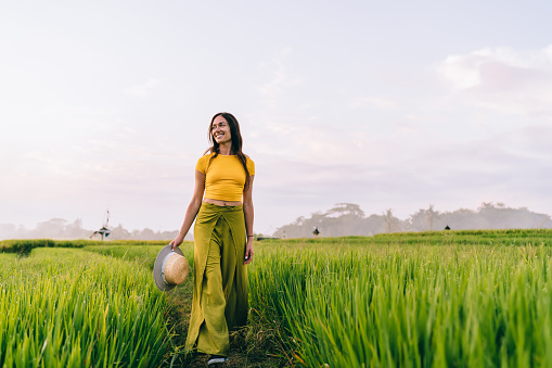 Full body of happy young woman in casual clothing walking through rice field in Philippines and looking into distance with toothy smile