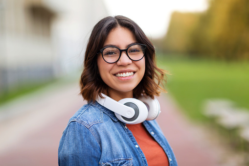 Portrait Of Young Arab Student Female In Stylish Eyeglasses Standing Outdoors, Happy Middle Eastern Woman With Wireless Headphones Posing Outside, Looking And Smiling At Camera, Copy Space