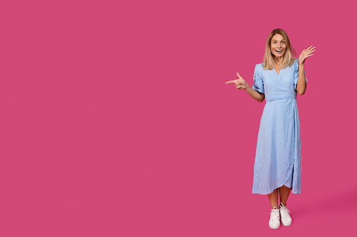 Cheerful shocked middle aged caucasian female blonde in dress pointing finger at empty space, isolated on pink background, studio, full length. Big surprise, great news, sale, advertising and offer