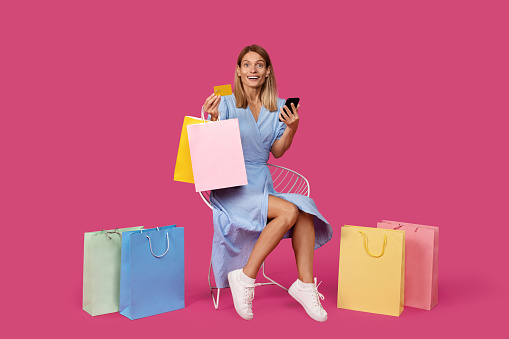 Glad shocked adult caucasian female blonde in dress with bags with purchases shows credit card and smartphone, isolated on pink background, studio. Ad and offer, online app for shopping and cashback