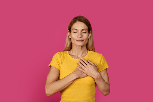 Happy calm pretty adult caucasian blonde woman in yellow t-shirt with closed eyes presses her hands to chest, heart, isolated on pink background, studio. People emotions, gratitude, thanks and love