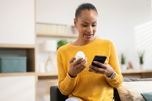 Woman holding medicine searching information online by smartphone sitting on sofa in living room at home.