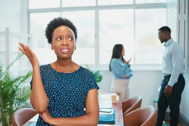 Photo of Young Black woman looks upset while colleagues talk behind her back office drama