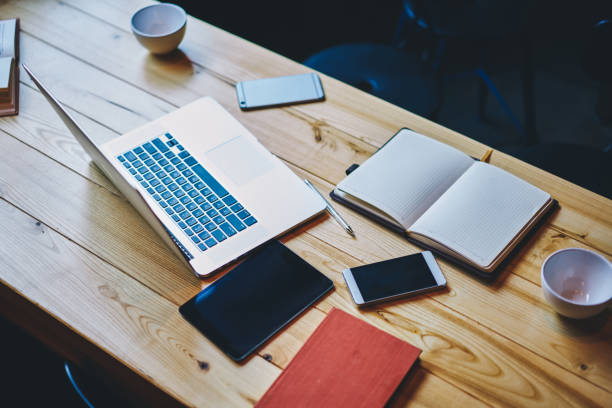 From above of wooden table with laptop and smartphone left by workers with notebooks and empty cups in modern workspace