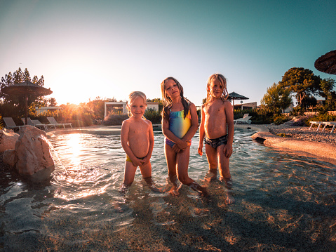 Caucasian siblings standing by hotel pool. Sunset shot of two males and one female child looking at camera.