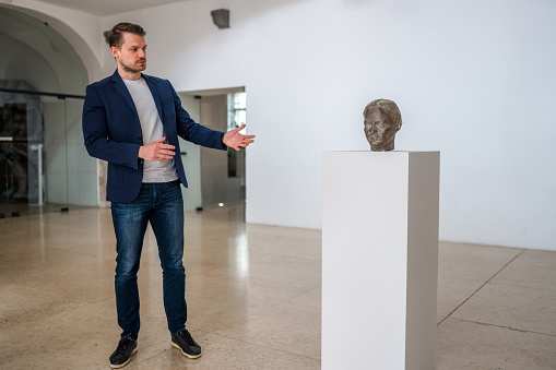 Mid adult Caucasian man, dressed casually, showing the bronze head on a white stand, explaining the details of the artwork. Full length image. Empty walls in the  background.