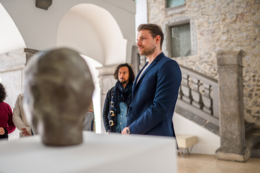 A group of people at a museum gallery, listening to a handsome Caucasian male expert standing next to the exhibited object. Waist up image, side view.  Natural light.