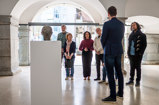 Full length image of a male art professor with a mixed group of pupils, all listening to him giving detailed explanation of the exhibited art object. Back view of the man. Bright light in the background.