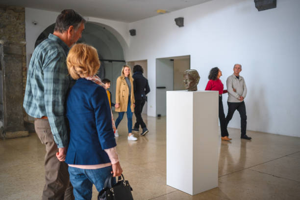 Back view of an older couple looking at an object on a stand, 3/4 length shot. Various people passing by in the background.