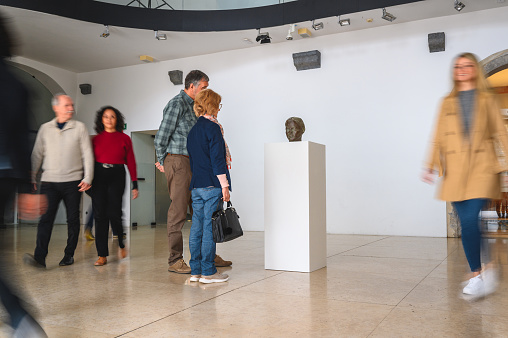 Caucasian female and her grey-haired husband in the center of an exhibition room. Side view of them looking at the object on a pedestal with other visitors blurred nearby. Full length image.