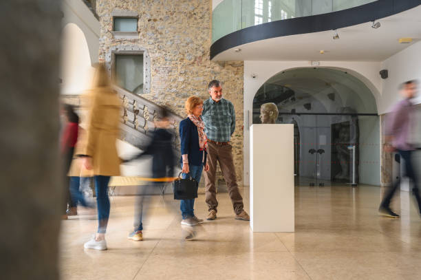 Retired Caucasian married couple on a gallery visit. A bronze head on a white pedestal in center of the room. Focus on the couple  closely observing it while other passers-by are  blurred. Full length shot.