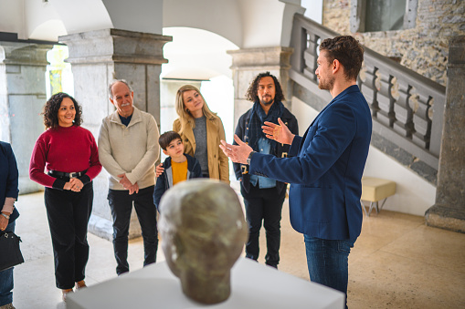 A varied group of members of an art course listening to a handsome male art teacher explaining the process of making a bronze head, exhibited on a stand. 3/4 length image.