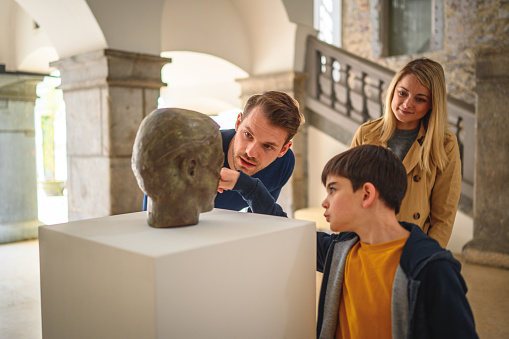 A close up of a bronze head of a man in a museum. The head is put on the white pedestal in the history museum lobby. The lobby is bright and modern.