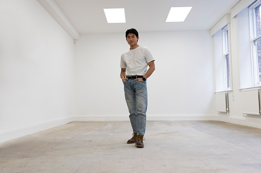 A wide-angle shot of a male Chinese man standing in an office space at his place of work. He is standing with his hands in his pockets and looking at the camera with a confident attitude.