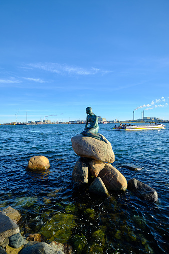 Copenhagen, Denmark - February  4, 2023: Statue of the Little Mermaid in Copenhagen with Tourist boat and Incinerator in the Background, some people visible on the boat.