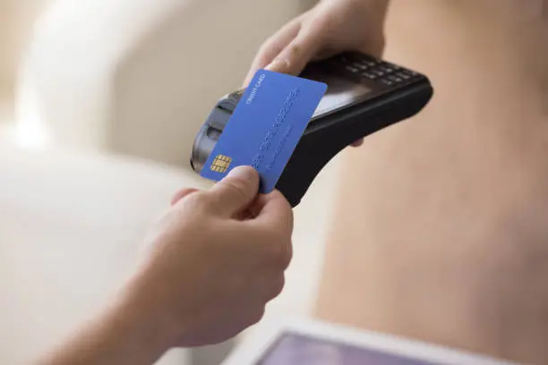 Hand of bank cardholder paying bill in cafe, applying blue credit card with chip at wireless payment terminal held by waitress, using electronic transaction banking technology. Clopped shot, close up